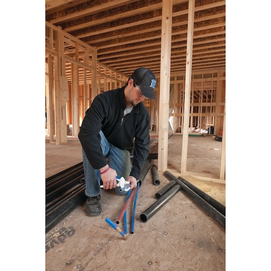 Photo of a person kneeling in a wood-framed construction site using a LENOX pipe cutter, SKU 12123R1, on blue and red pipes.