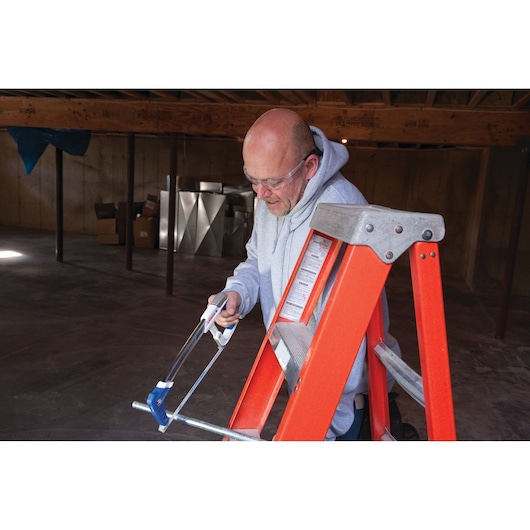 Photo of LENOX 12132HT50 hacksaw cutting metal pipe on orange ladder in unfinished room.