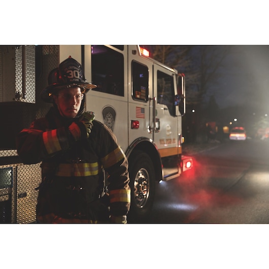 A firefighter in full uniform stands next to a fire truck on a smoky street at night. The firefighter's face is blurred for privacy.
