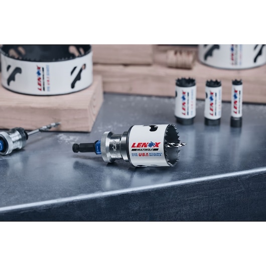 A close-up image of several Lenox hole saw drill bits displayed on a metal surface, with wood blocks in the background. The focus is on the main drill bit in the front, showing the Lenox branding and USA origin.