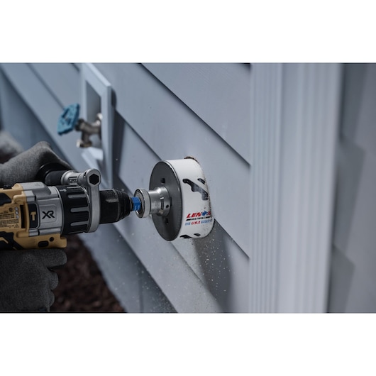LENOX 3006666L hole saw attached to drill cutting through exterior siding, close-up side view.