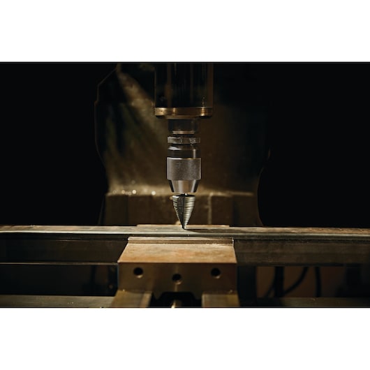 LENOX step drill bit 30912VB12 mounted on drill press boring into metal bar, close-up, dark background.