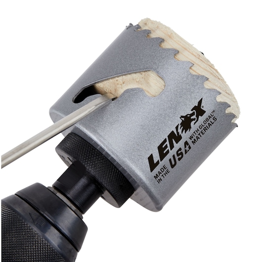 Close-up of a LENOX hole saw drill bit cutting through a piece of wood. The tool is attached to a drill, and the LENOX branding with 'Made in the USA with global materials' is visible on the side.
