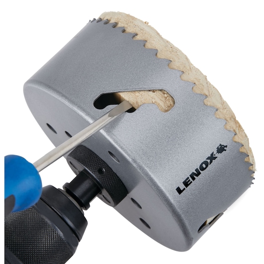 Close-up of a LENOX branded hole saw attached to a power drill, with a screwdriver removing a wooden plug from inside the saw.