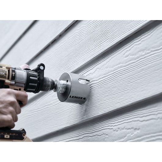 Photo of LENOX LXAH4358 hole saw attached to a drill cutting through gray exterior siding.