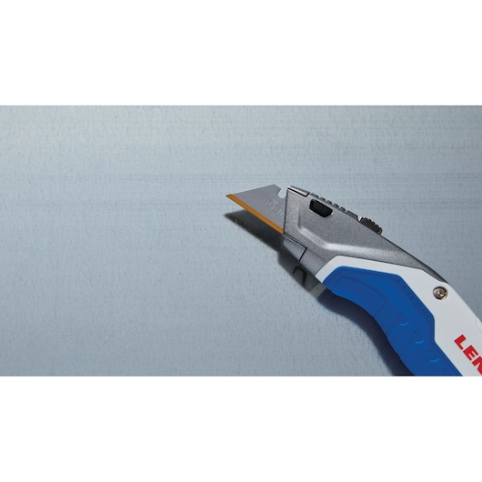 Photo of LENOX utility knife LXHT10599 with blue and white handle, partially visible, cutting a grey surface.