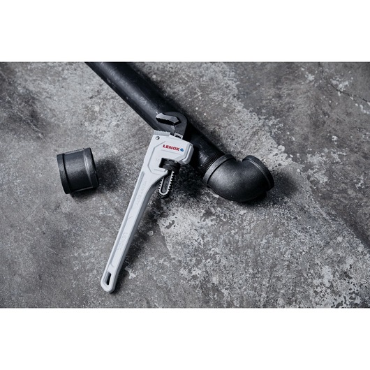 A LENOX pipe wrench placed on a black metal pipe and fittings, set on a grey concrete surface.